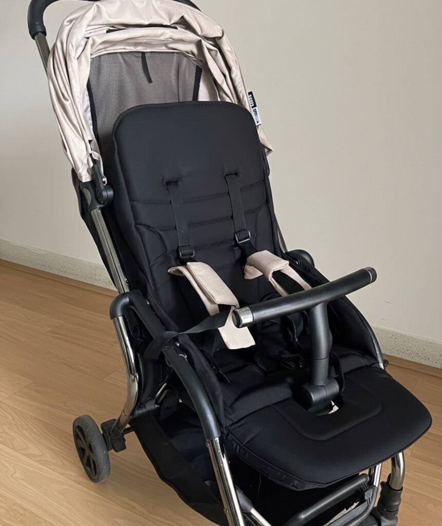 좌판과 등받이는 검정색, 차양막은 베이지색인 휴대용 유모차가 방 안에 놓여있다